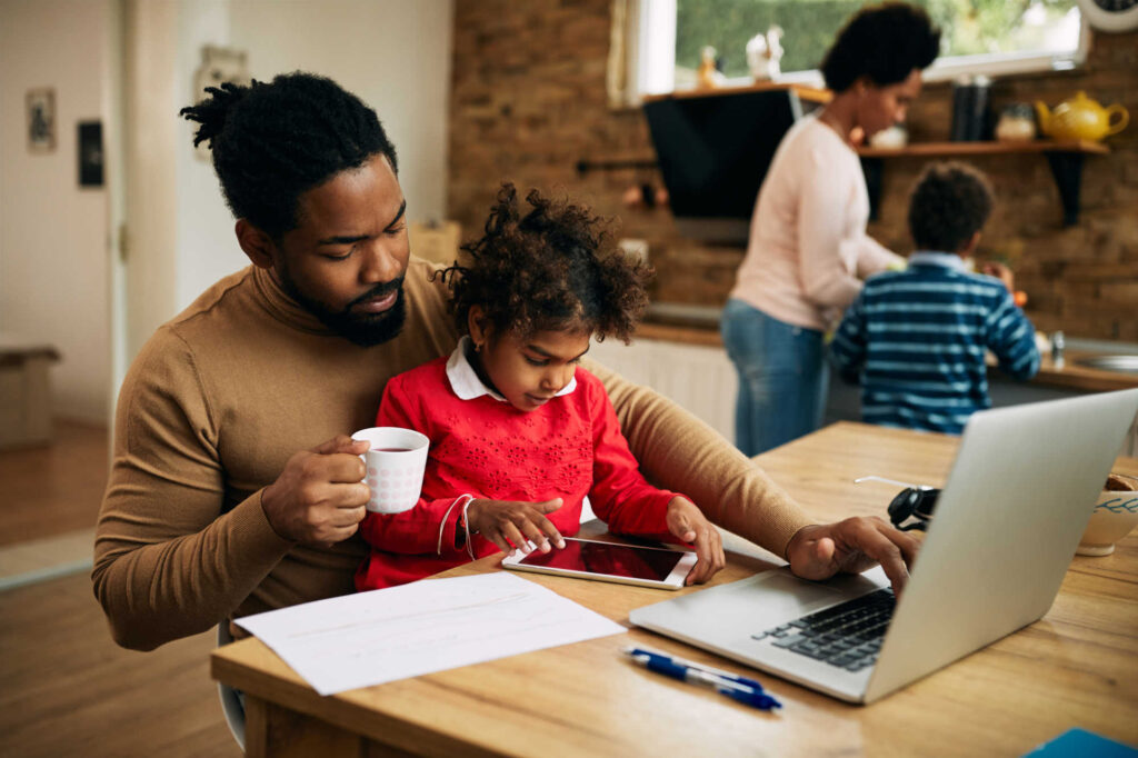 picture of a black family in their dining area. A young child is sitting on the lap of the father while he works on a laptop and the mother and other child do food prep in the background.
